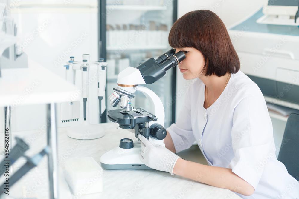 Female lab scientist researcher technician worker in gloves sits next to a microscope in laboratory Female lab scientist researcher technician worker in gloves sits next to a microscope in laboratory