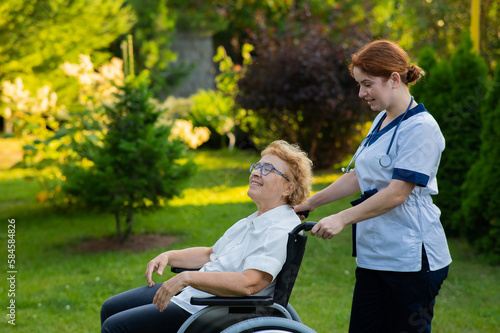 Caucasian female doctor walks with an elderly patient in a wheelchair in the park. Nurse accompanies an old woman on a walk outdoors.  © Михаил Решетников