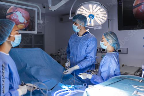 Back view of surgeons team looks at monitors while preforming operation in hospital operating theater, male surgeon operating patient working with surgical laparoscopy instruments.