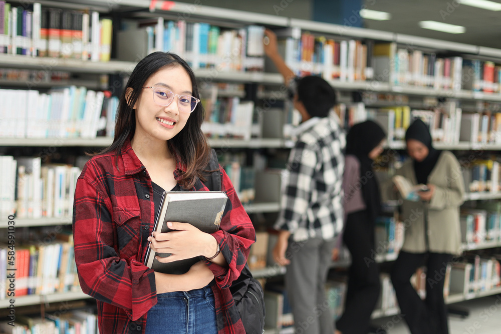 Pretty smart young Asian student standing confidently and smiling to the camera, hold book, against blurry background of diverse student at library. Pretty smart young Asian student standing confidently and smiling to the camera, hold book, against blurry background of diverse student at library.