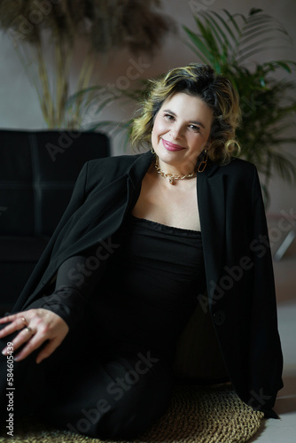 A smiling woman in a black suit is sitting on a rug in the room.