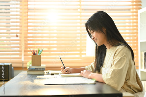 Smiling asian teen girl making notes, ding essay homework assignment at home. Learning and education concept