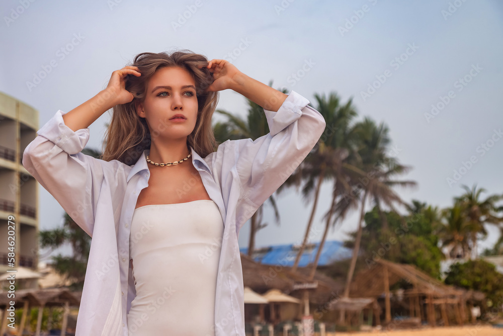 Portrait happy pretty young woman in white shirt posing on sandy beach at palm trees background. Relaxing and enjoyable vacation in tropical coastline. Travel vacation holiday concept. Copy text space