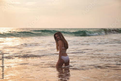 Back view of young slim woman in white swimsuit posing in water at sea background  looking at camera. Relaxing and enjoyable vacation on tropics. Travel vacation holiday concept. Copy ad text space
