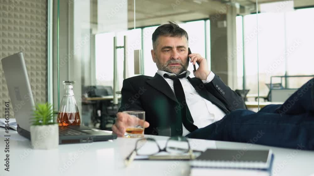 Senior businessman drinking from stress in office putting legs on table and talking on the phone. Alcoholism, drinking concept Senior businessman drinking from stress in office putting legs on table and talking on the phone. Alcoholism, drinking concept