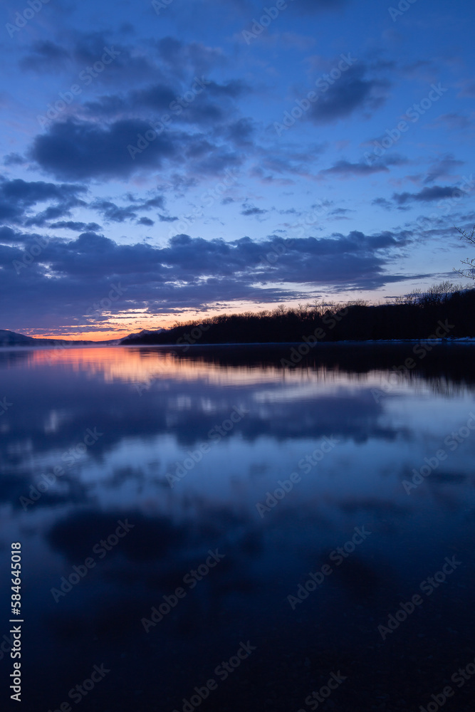 雲の浮かぶ夜明けの空と湖畔の森を湖面に映す湖。日本の北海道の屈斜路湖。