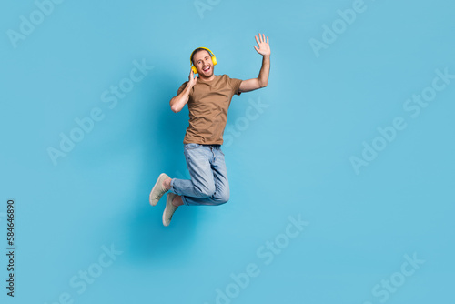 Full length photo of cheerful dreamy man wear beige t-shirt jumping high listening songs empty space isolated blue color background