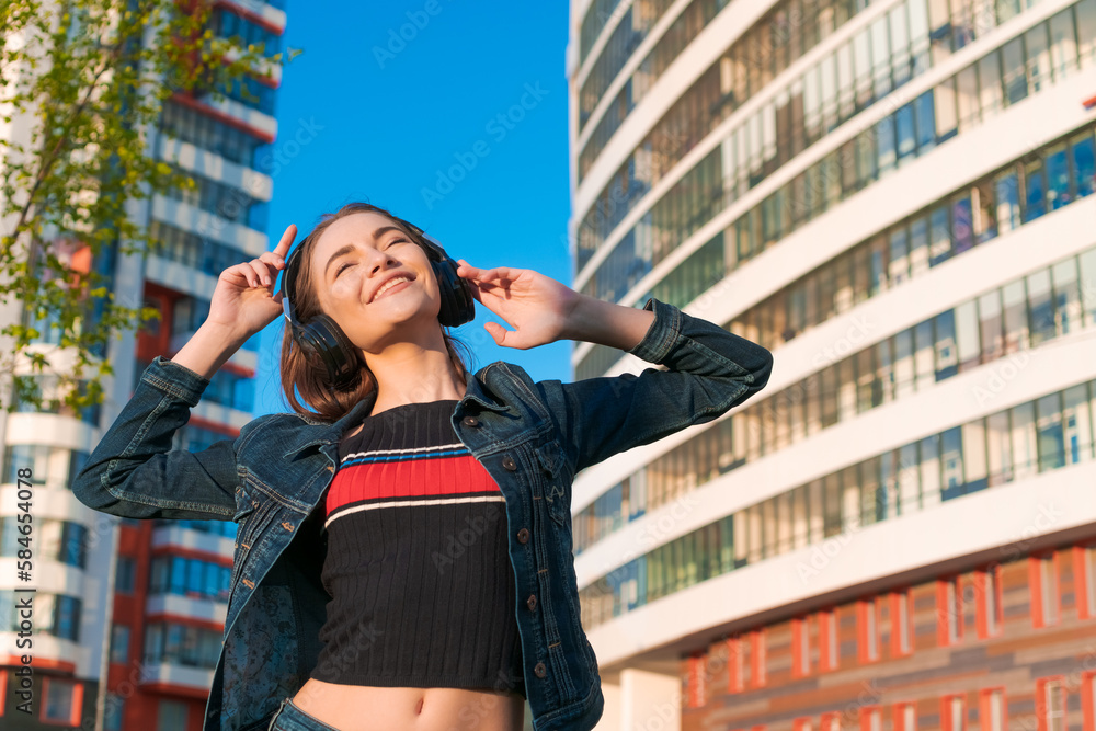 Young energetic cheerful caucasian woman wearing headphones. Dressed in a denim jacket, enjoying the spring sun. People urban youth lifestyle.