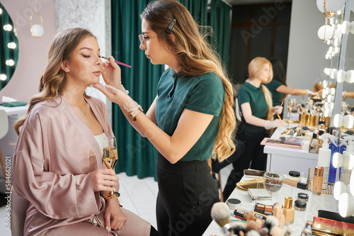 Beauty specialist applying eyeshadow on client eyelid in visage studio. Young woman sitting at dressing table and holding glass of champagne while makeup artist doing professional makeup. photo