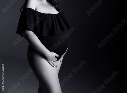 Studio pregnancy shoot. A pregnant woman in a black bodysuit holds her belly. A pregnant woman in a black bodysuit stands on a black background with a place for text. Pregnant woman in black dress.