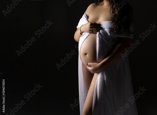 A pregnant woman in a white dress on a gray background with a place for text. Belly of a pregnant woman in close-up. Studio pregnancy photo shoot.