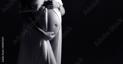 Pregnant woman in a white dress on a black background. Pregnancy studio shooting. Person in a dress. Copy space. Black and white photo. Pregnant silhouette.