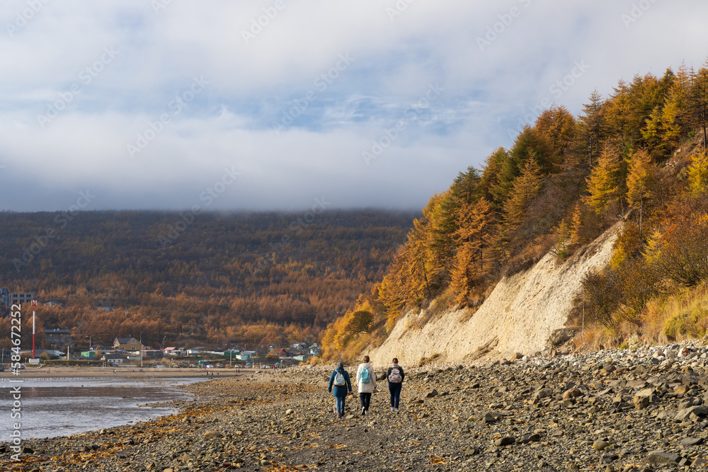 Beautiful autumn landscape. View of the seashore at low tide. Forest on the coast. Seaside village in the distance. Three women with backpacks walk along the shore. Low clouds over the mountains.