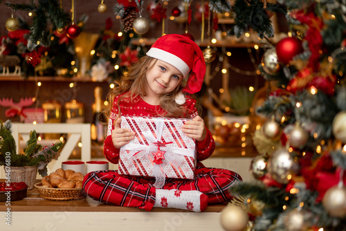 a little cute girl child in a red sweater and a Santa Claus hat in a dark kitchen with a Christmas tree rejoices with gifts and waits for the new year or Christmas