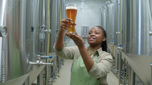 Medium arc shot of African American female brewery technologist pouring glass of freshly brewed lager from fermenter tank, lifting up, checking texture and carbonation, and smiling with satisfaction