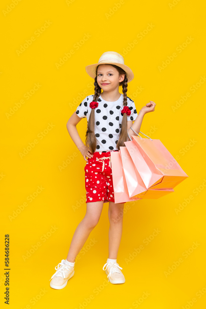 A little girl holds shopping bags. A child in a summer hat and shorts is engaged in children's shopping. A happy child in full growth on a yellow isolated background. A little girl holds shopping bags. A child in a summer hat and shorts is engaged in children's shopping. A happy child in full growth on a yellow isolated background.