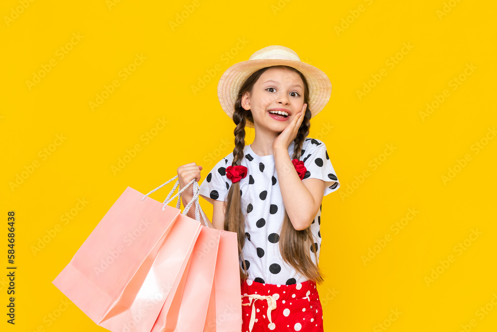 A child with paper bags from the store. Summer shopping for children. A beautiful little girl in a hat is smiling broadly. Yellow isolated background. A child with paper bags from the store. Summer shopping for children. A beautiful little girl in a hat is smiling broadly. Yellow isolated background.