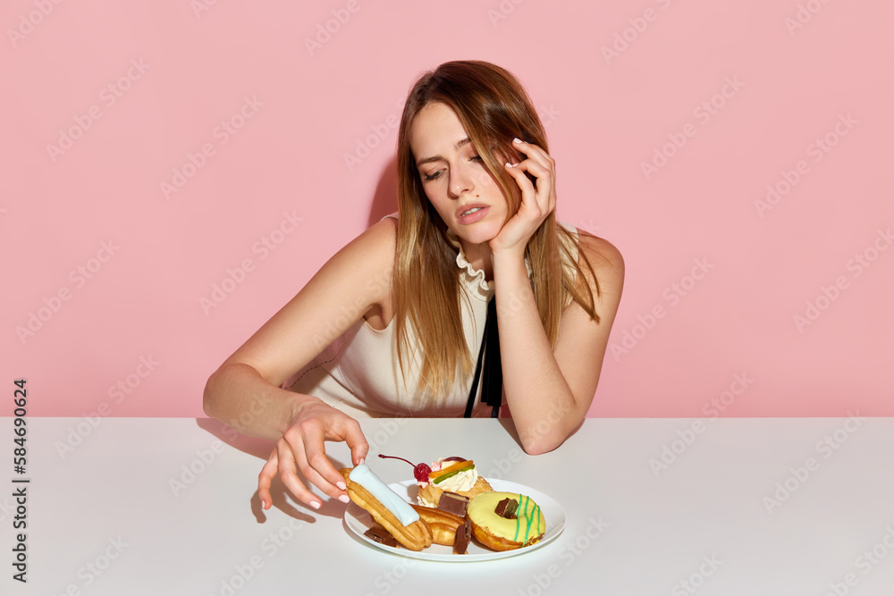 Adorable girl sitting at table and taking ecler with sad facial expression over pink background Adorable girl sitting at table and taking ecler with sad facial expression over pink background