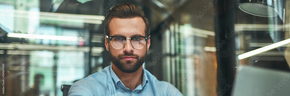 Confidence. Young bearded businessman in eyeglasses and formal wear looking at camera and keeping arms crossed while sitting in modern office Confidence. Young bearded businessman in eyeglasses and formal wear looking at camera and keeping arms crossed while sitting in modern office