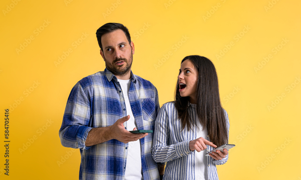 Obraz premium Surprised girlfriend with mouth open using mobile phone and looking at rude boyfriend rolling his eyes in irritation and ignoring her on isolated yellow background