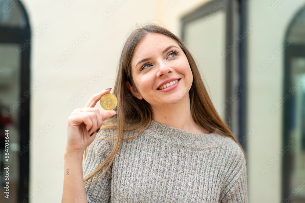Young pretty blonde woman holding a Bitcoin at outdoors looking up while smiling
