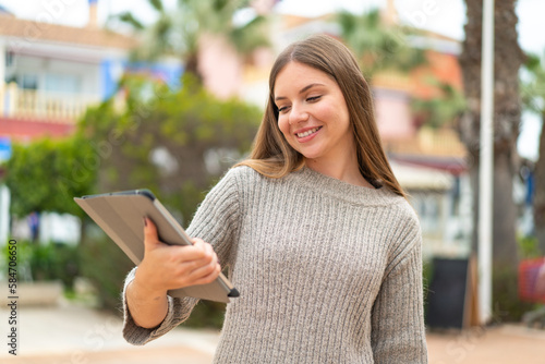 Young pretty blonde woman holding a tablet at outdoors with happy expression