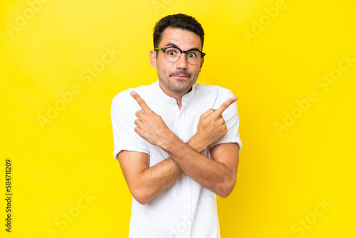 Young handsome man over isolated yellow background pointing to the laterals having doubts
