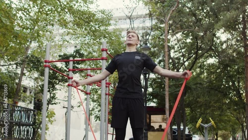 Young caucasian male athlete tensely performing lateral raises with a flat resistance band in an outdoor workout area. Sportsman doing exercises for shoulders