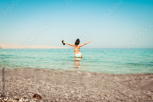 Young carefree Caucasian woman tourist dance on the beach in turquoise water alone with hands up hold slippers on summer vacation beach holidays. Solo travel adventure wanderlust