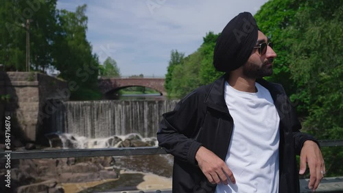 Punjabi Sikh Man In Sunglasses And Dastar Turban Standing On The Bridge With Vanhankaupunkoski, Old Town Waterfall In Helsinki, Finland. - medium photo