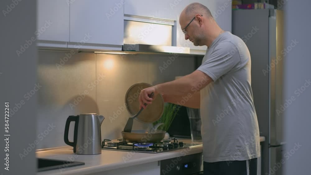 Handsome man in casual clothes panning meat on stove at kitchen. Husband preparing dinner at home. Hobby, preparing food home, modern interior, cookery concept Handsome man in casual clothes panning meat on stove at kitchen. Husband preparing dinner at home. Hobby, preparing food home, modern interior, cookery concept