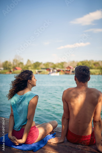 Man and woman sitting by water on wooden jetty. Rear view of couple. Woman stretching. Togetherness, holiday, lifestyle concept.