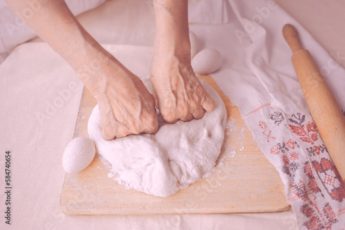 Elderly woman hands rolling out dough in flour with rolling pin in her home kitchen. Close-up of overworked old woman hands making home made baking, pastry and cookery.