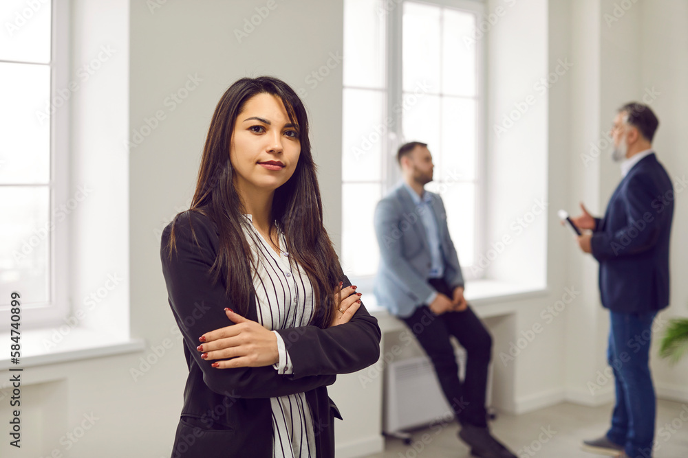 Portrait of a young business woman or office employee at work. Beautiful young female in a suit standing with her arms crossed in the office, with her colleagues in the background Portrait of a young business woman or office employee at work. Beautiful young female in a suit standing with her arms crossed in the office, with her colleagues in the background