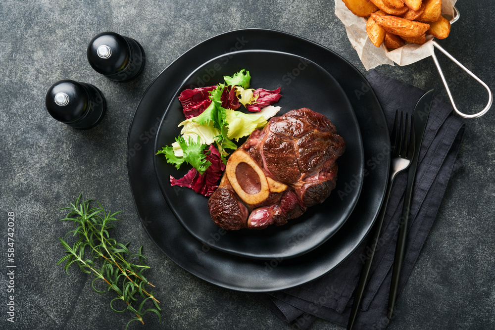 Steaks Osso Buco. Sliced beef grilled Osso Buco or porterhouse meat steak with garnished with salad and french fries on black marble board on old wooden background. Top view. Mock up. Steaks Osso Buco. Sliced beef grilled Osso Buco or porterhouse meat steak with garnished with salad and french fries on black marble board on old wooden background. Top view. Mock up.