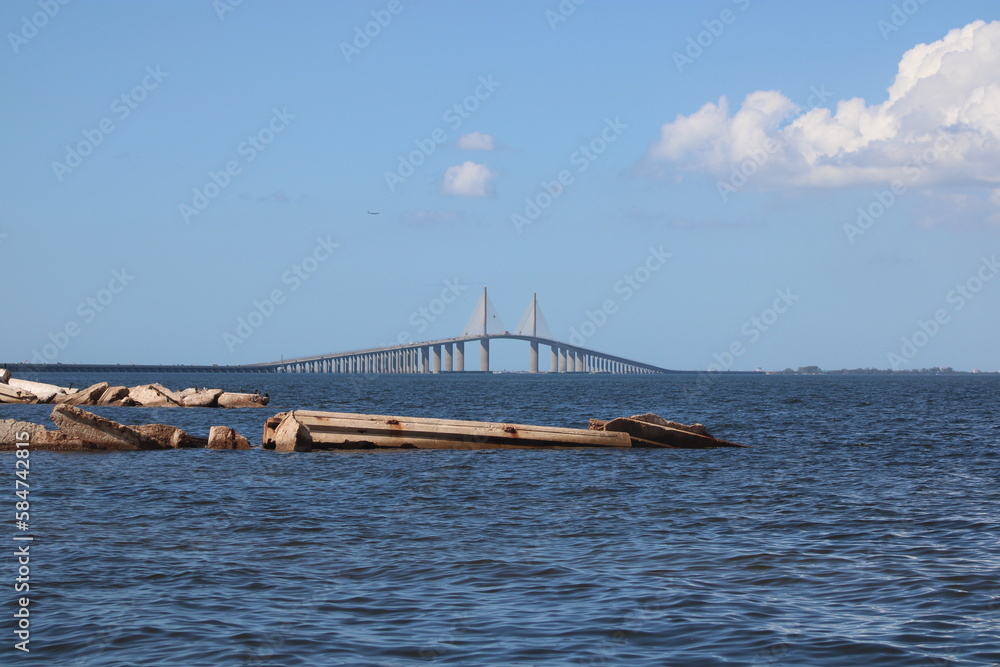 Obraz premium A broken jetty just before the Sunshine Skyway Bridge to St. Petersburg Florida. the jetty and pier are in focus with the bridge in the background. Deep blue Tampa bay water and blue sky with clouds