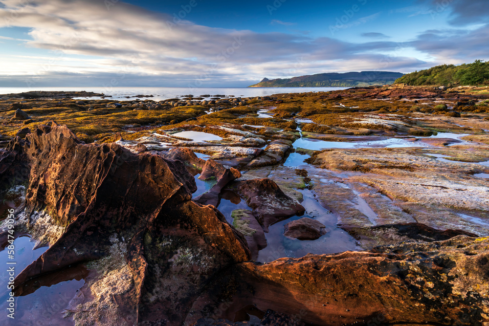 Sandstone patterns and finned bedrock along the rocky coastline at Pirate's Cove near Corrie, Isle of Arran, Scotland, with the peak of Holy Island in the distance.