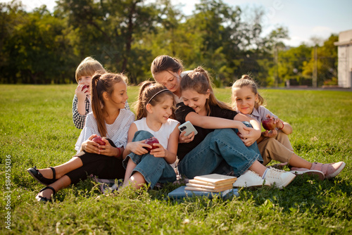 children play in smartphone or watch video together sitting on green grass in park or school yard