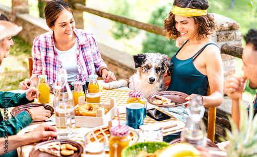 Young friends on healthy pic nic break fast with cute puppy at countryside farm house - Unplug life style concept with happy people having fun together out side at garden picnic party - Focus on dog