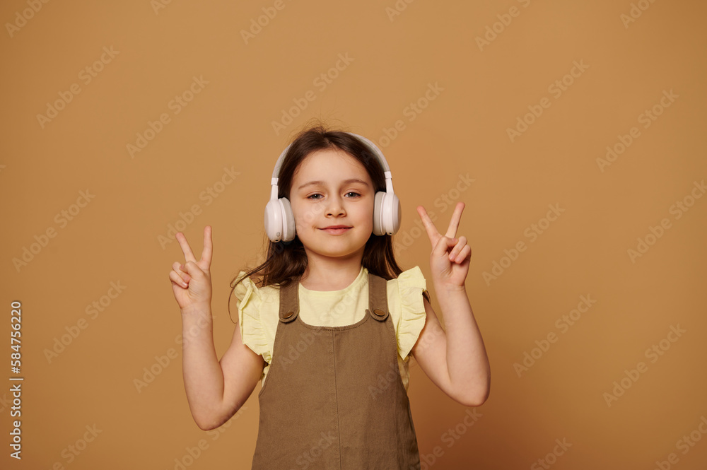 Caucasian cheery little child girl 5-6 years old, wearing wireless headphones, showing peace sign with her fingers, expressing positive emotions, smiling looking at camera, isolated beige background Caucasian cheery little child girl 5-6 years old, wearing wireless headphones, showing peace sign with her fingers, expressing positive emotions, smiling looking at camera, isolated beige background