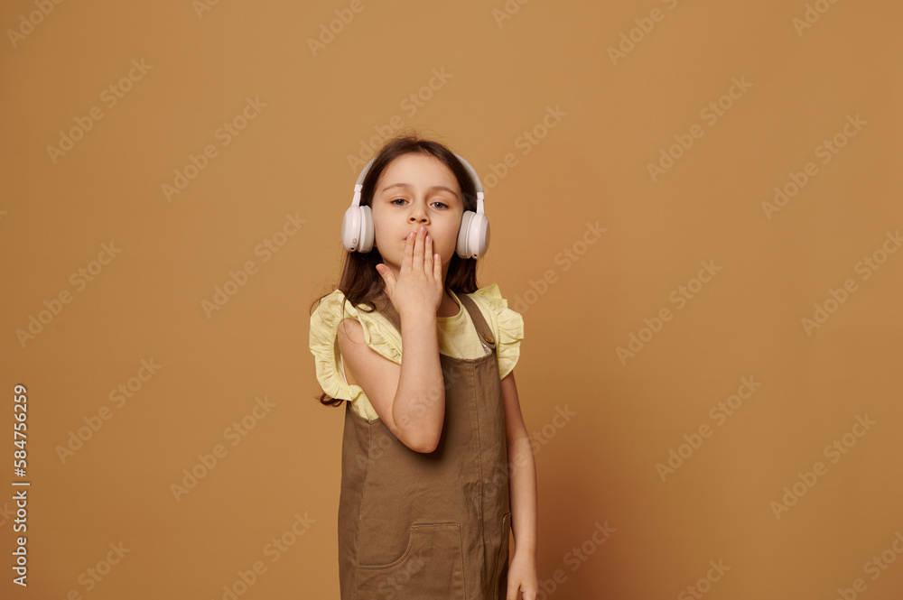 Obraz premium Caucasian adorable little child girl dressed in stylish khaki dress, listening to music on wireless headphones, blowing out and sending air kisses looking at camera, isolated over beige background.