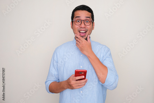 Adult Asian man showing wow face expression when holding a mobile phone photo