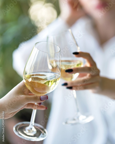 Glasses of white wine in hands of women. Two women drink white wine. Cheers, clinking of glasses. Holiday celebration. Meeting and chatting with friends. Front view. Soft focus. Close-up. 
