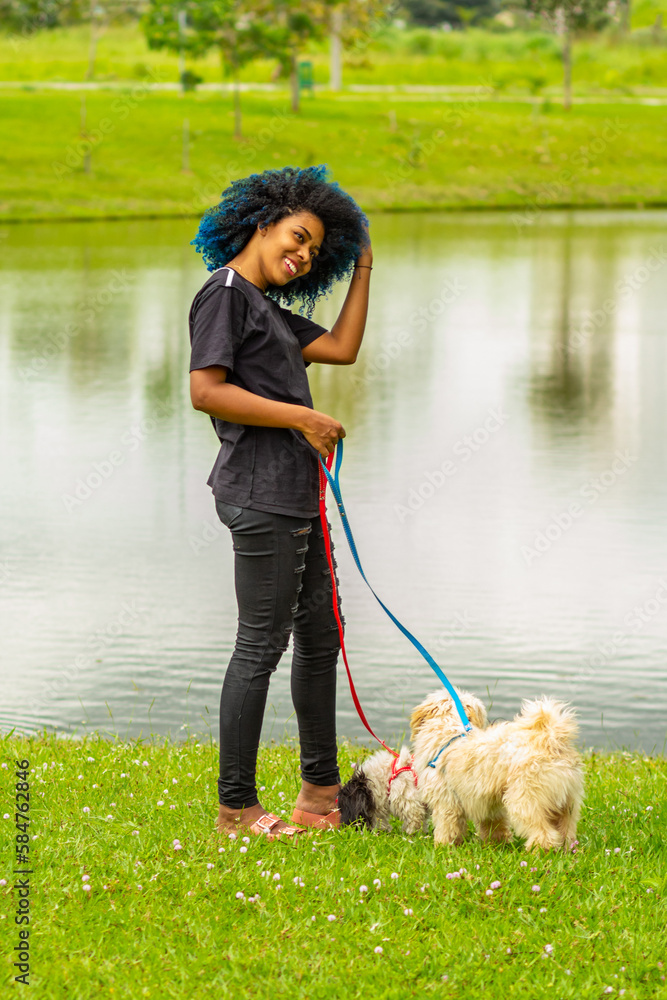 Uma jovem mulher negra, com cabelos afro pintados de azul, passeando com seus cachorros no gramado ao lado de um lago no parque. Uma jovem mulher negra, com cabelos afro pintados de azul, passeando com seus cachorros no gramado ao lado de um lago no parque.