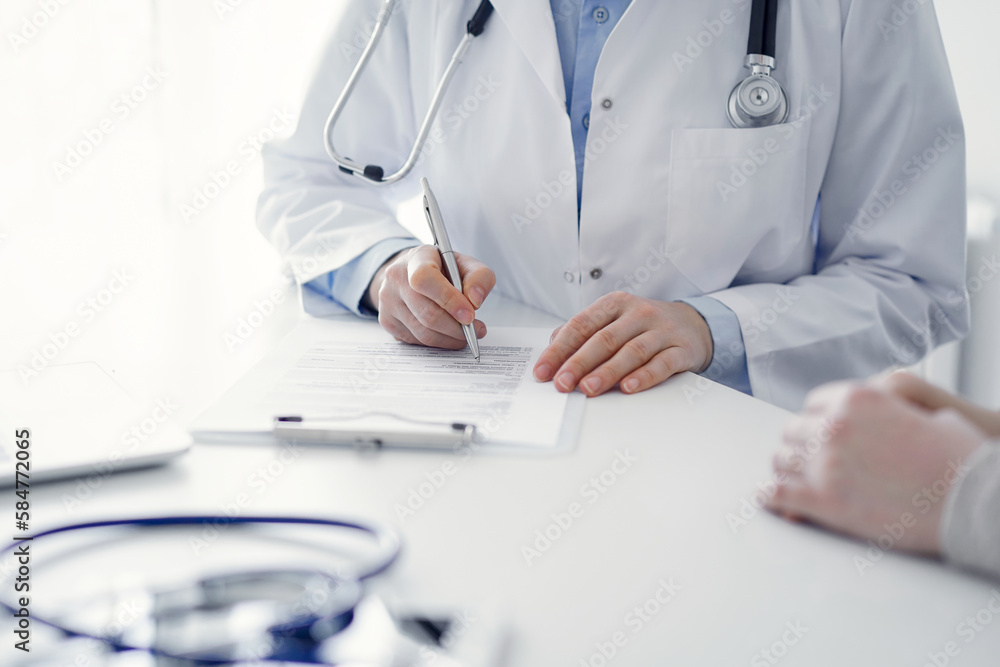 Doctor and patient sitting at the table in clinic. The focus is on female physician's hands filling up the medication history record form or checklist, close up. Medicine concept Doctor and patient sitting at the table in clinic. The focus is on female physician's hands filling up the medication history record form or checklist, close up. Medicine concept