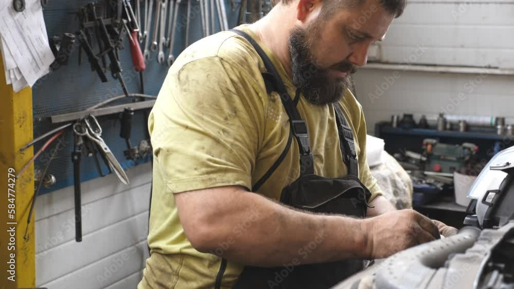 Profile of auto mechanic in uniform works with car at garage. Professional bearded repairman fixing vehicle at workshop or service. Concept of automobile maintenance and diagnostic. Close up Slow mo Profile of auto mechanic in uniform works with car at garage. Professional bearded repairman fixing vehicle at workshop or service. Concept of automobile maintenance and diagnostic. Close up Slow mo