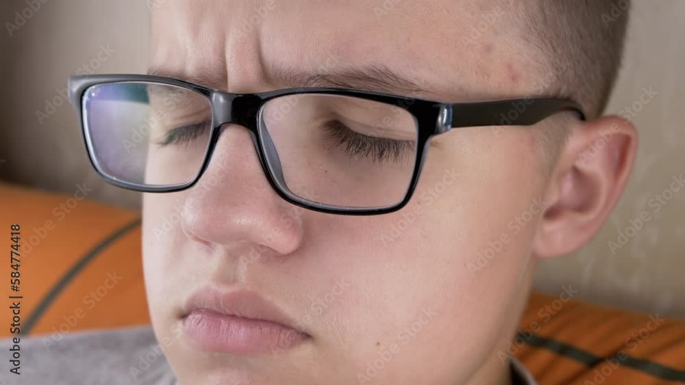 Close-up Face of an Offended Serious Schoolboy with Glasses, with Full Lips. Frowning, tired, sad teenager with blinking eyes, long eyelashes looking at a TV in bed. Poor eyesight. Children emotions. Close-up Face of an Offended Serious Schoolboy with Glasses, with Full Lips. Frowning, tired, sad teenager with blinking eyes, long eyelashes looking at a TV in bed. Poor eyesight. Children emotions.