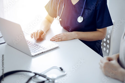 Doctor and patient sitting at the desk in clinic office. The focus is on unknown female physician's hands using a laptop computer, close up. Medicine concept