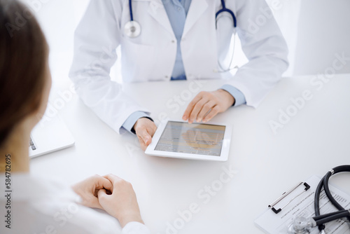 Doctor and patient sitting opposite each other at the desk in clinic. The focus is on female physician's hands pointing into tablet computer touchpad, close up. Medicine concept