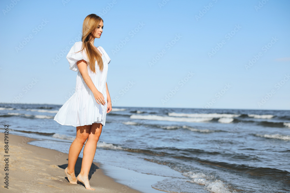 Happy smiling beautiful woman is walking on the ocean beach in a white summer dress